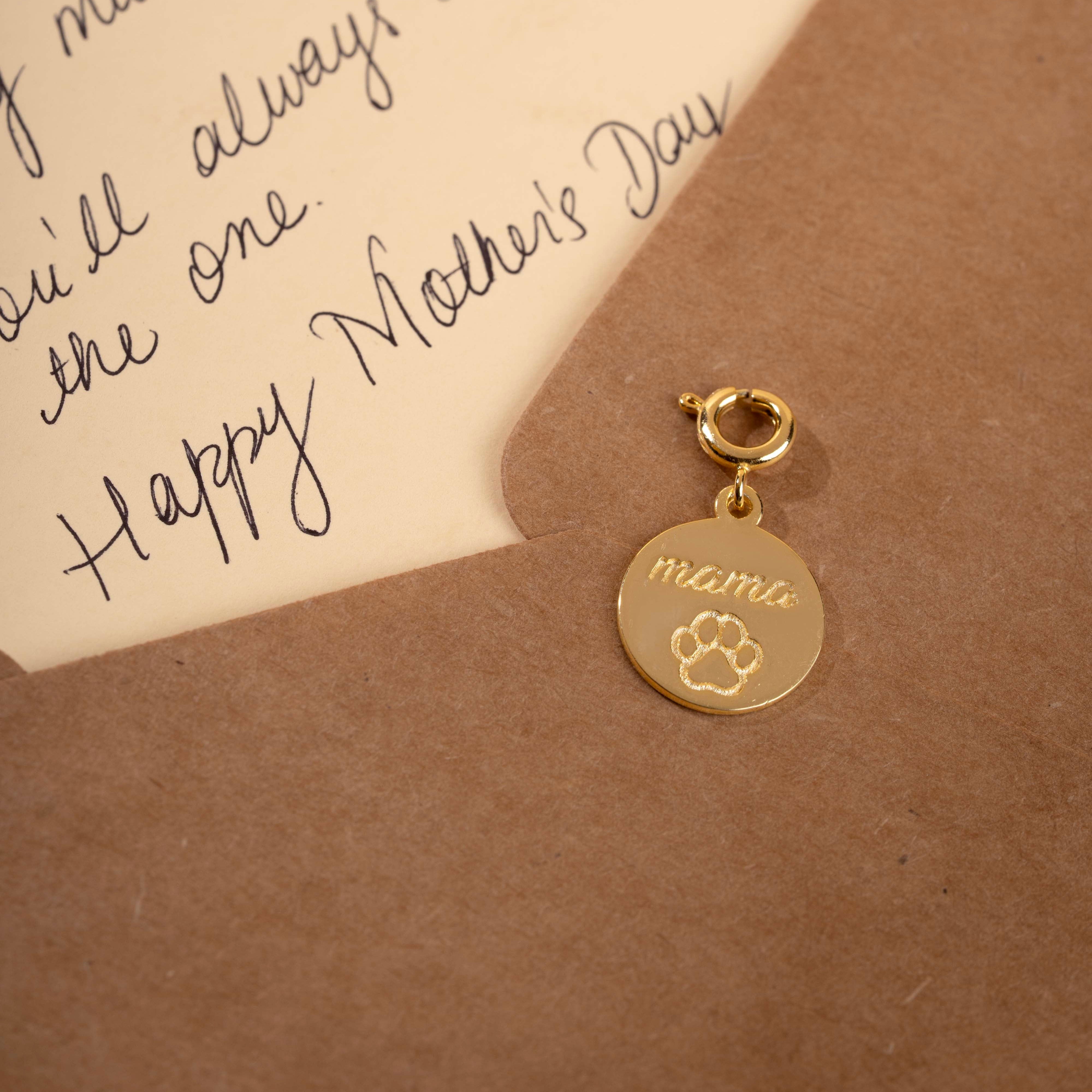 Gold charm with 'mama' and paw print on a brown envelope with handwritten text.